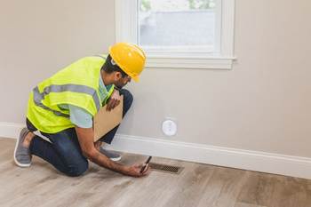 A man in a hard hat and safety vest inspects a floor vent during a home inspection. 