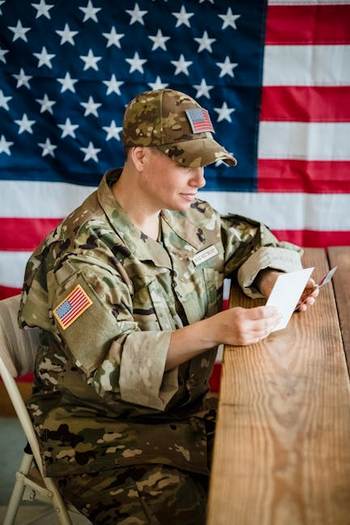 A national guardsman in uniform reads a letter at a table, with an American flag displayed nearby.  